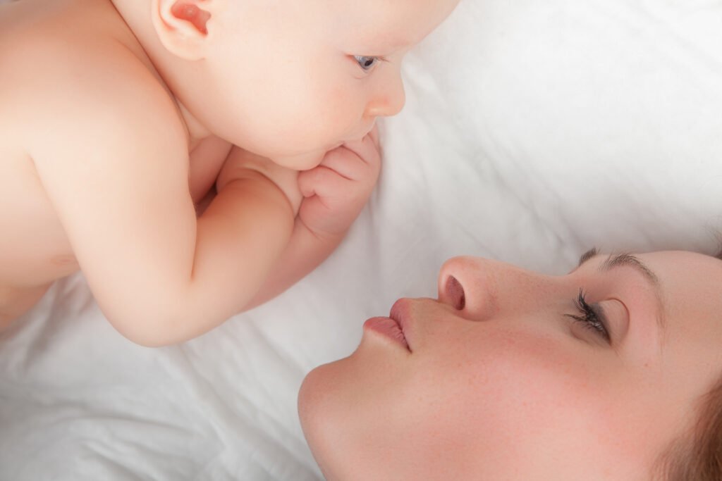 “Newborn baby lying close to mother, gazing into each other’s eyes while bonding on a white bed.”