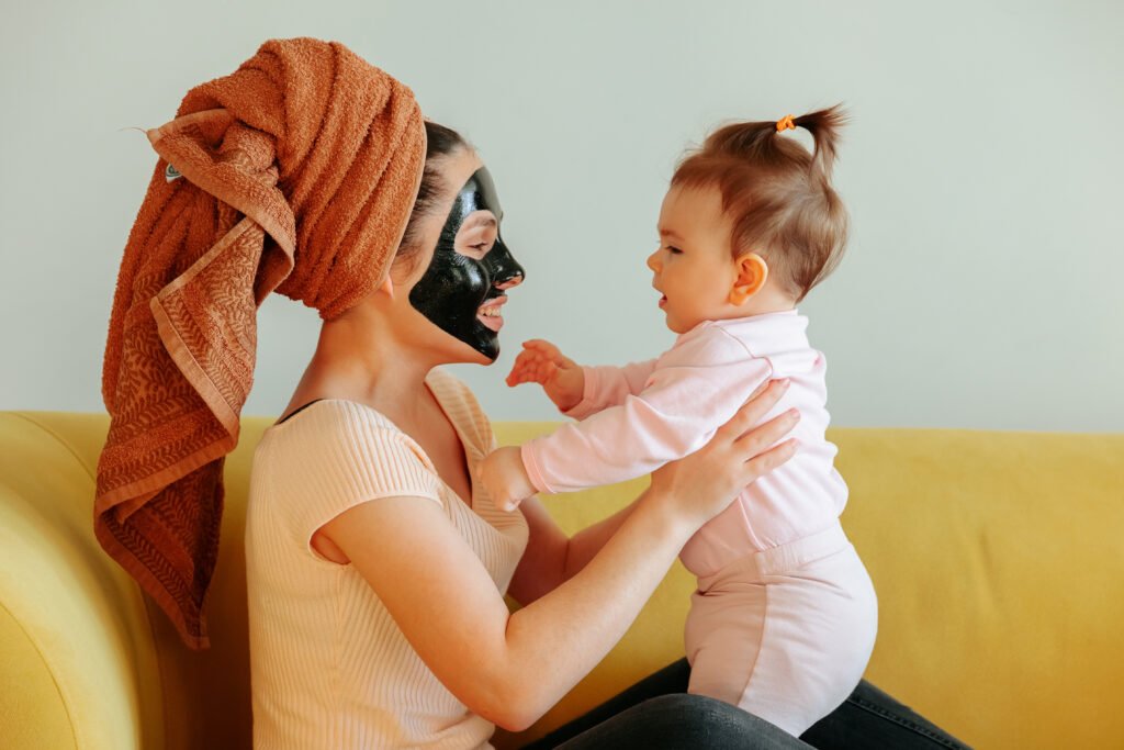 “Smiling mother with a black facial mask and towel on her head playing with her baby daughter at home.”
