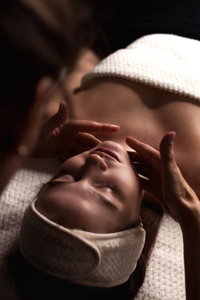 “Woman lying peacefully with eyes closed during a professional facial massage, wrapped in a spa towel.”